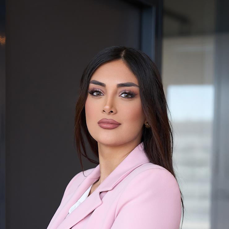 Frau mit langen dunklen Haaren in rosa Blazer steht mit verschränkten Armen vor einer Glastür.