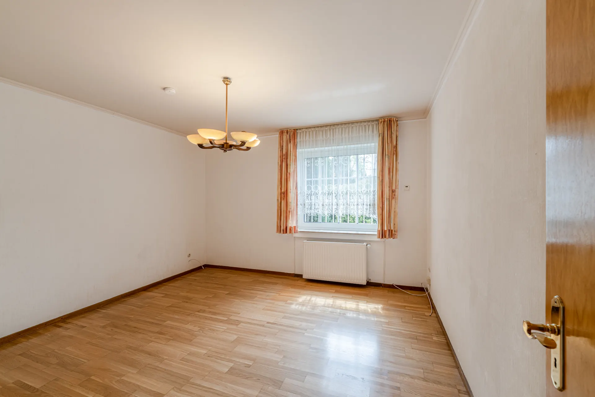 Leerer Raum mit Holzfußboden, Deckenlampe und Fenster mit Vorhängen.