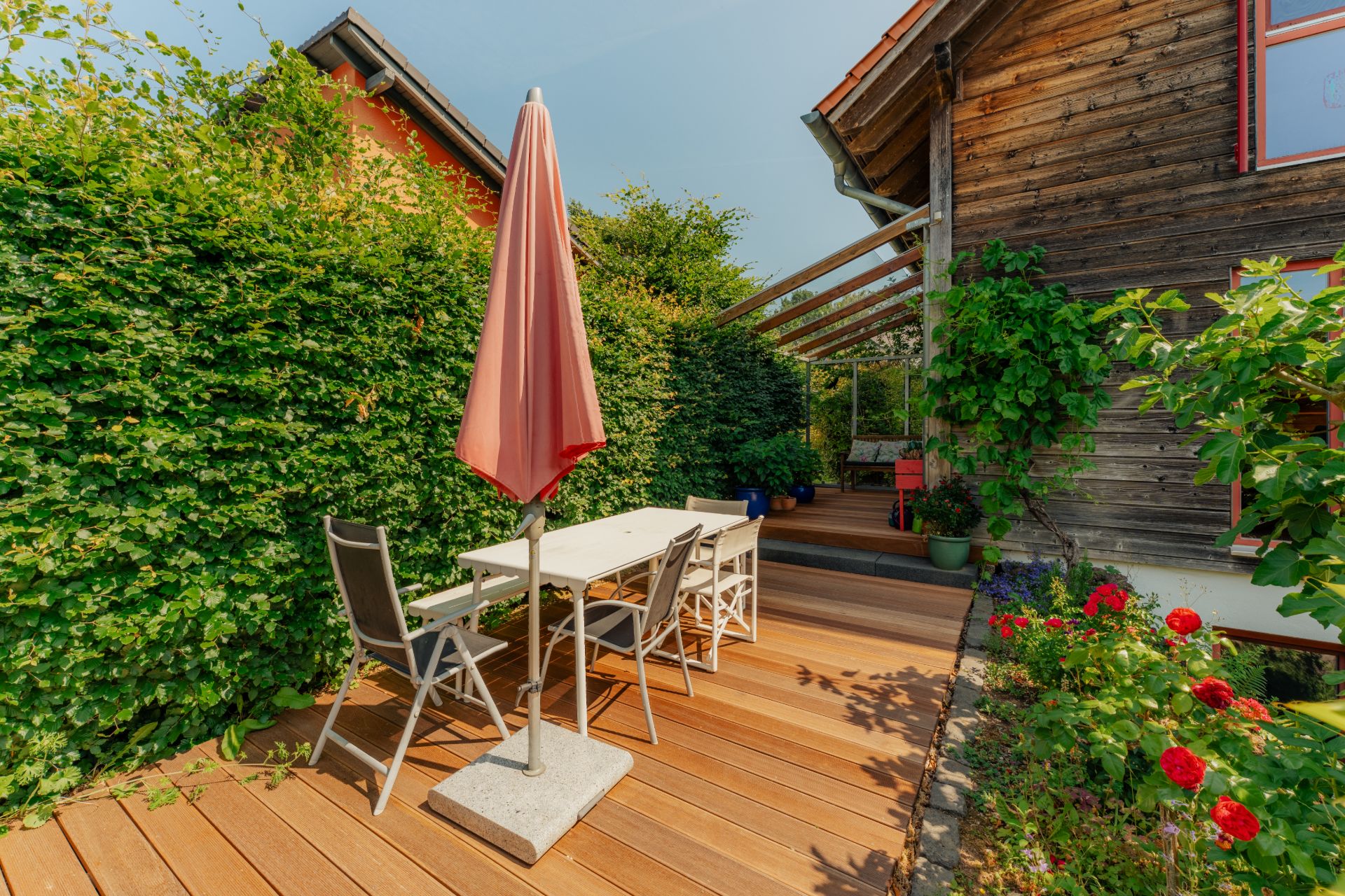 Holzterrasse mit Tisch, Stühlen, Sonnenschirm und umgebenden Pflanzen neben einem Haus.