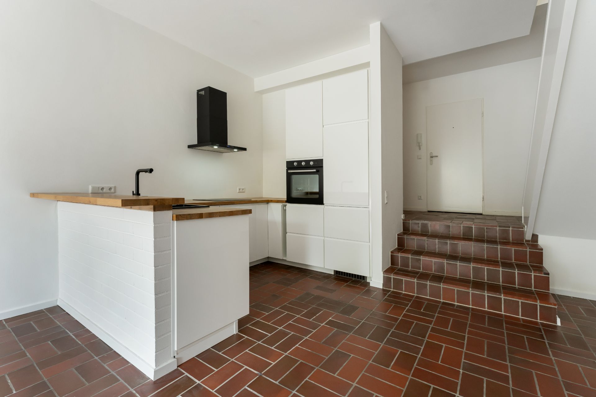 Moderne weiße Küche mit Holzarbeitsplatte und Backofen, daneben eine Treppe mit braunen Fliesen.