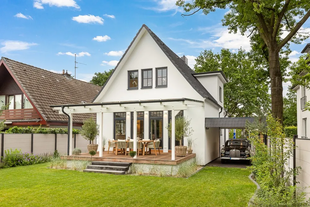 Weißes Einfamilienhaus mit Terrasse, Garten und Carport, umgeben von Bäumen und Nachbarhäusern.