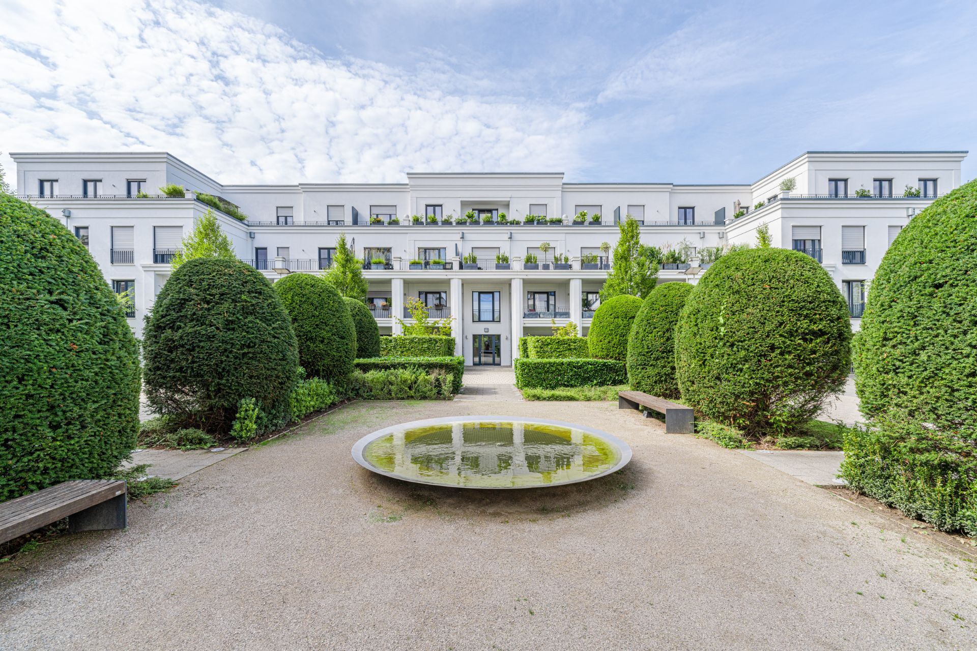 Moderner Wohnkomplex mit gepflegtem Garten, runden Büschen, Bänken und flachem Wasserbecken im Vordergrund.