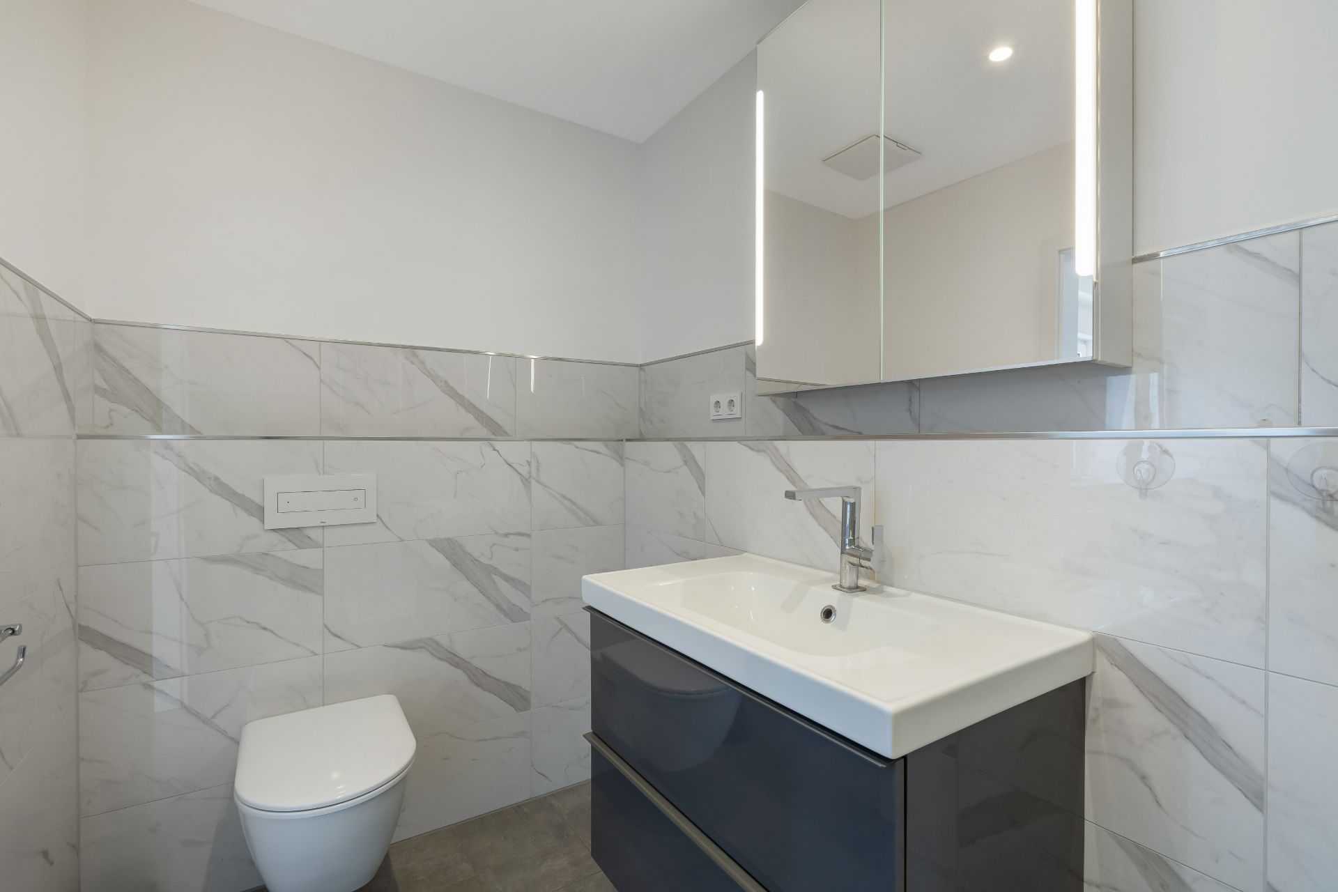 Modernes Badezimmer mit Wandtoilette, Waschbecken und beleuchtetem Spiegel.