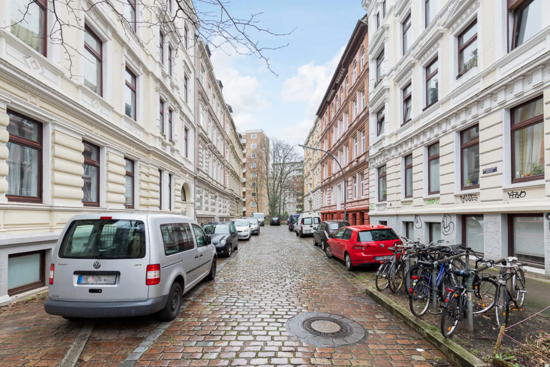 Gepflasterte Wohnstraße mit geparkten Autos und Fahrrädern vor Altbauten.