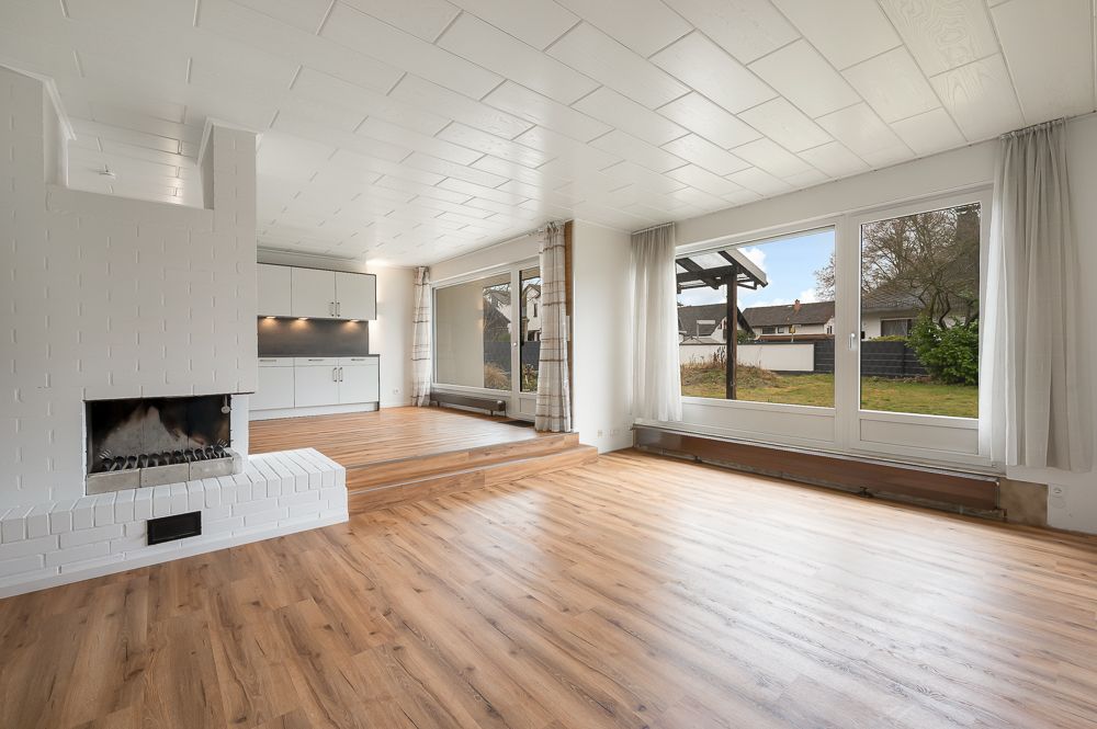 Helles Wohnzimmer mit Kamin, Holzfußboden, großer Fensterfront und offener Küche.