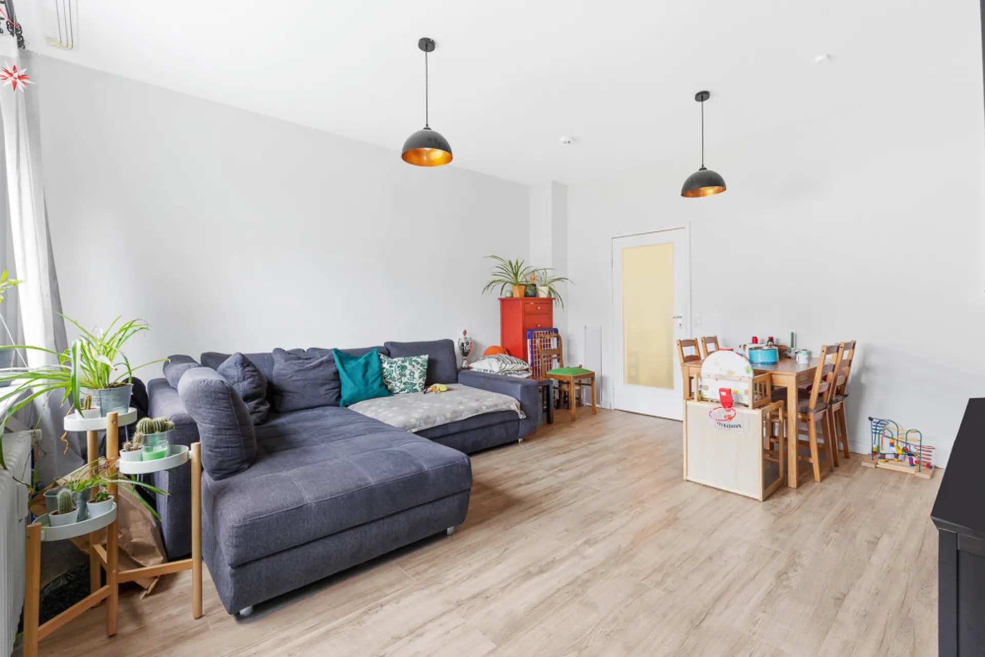 Wohnzimmer mit grauer Eckcouch, Esstisch, Spielzeug und mehreren grünen Pflanzen.