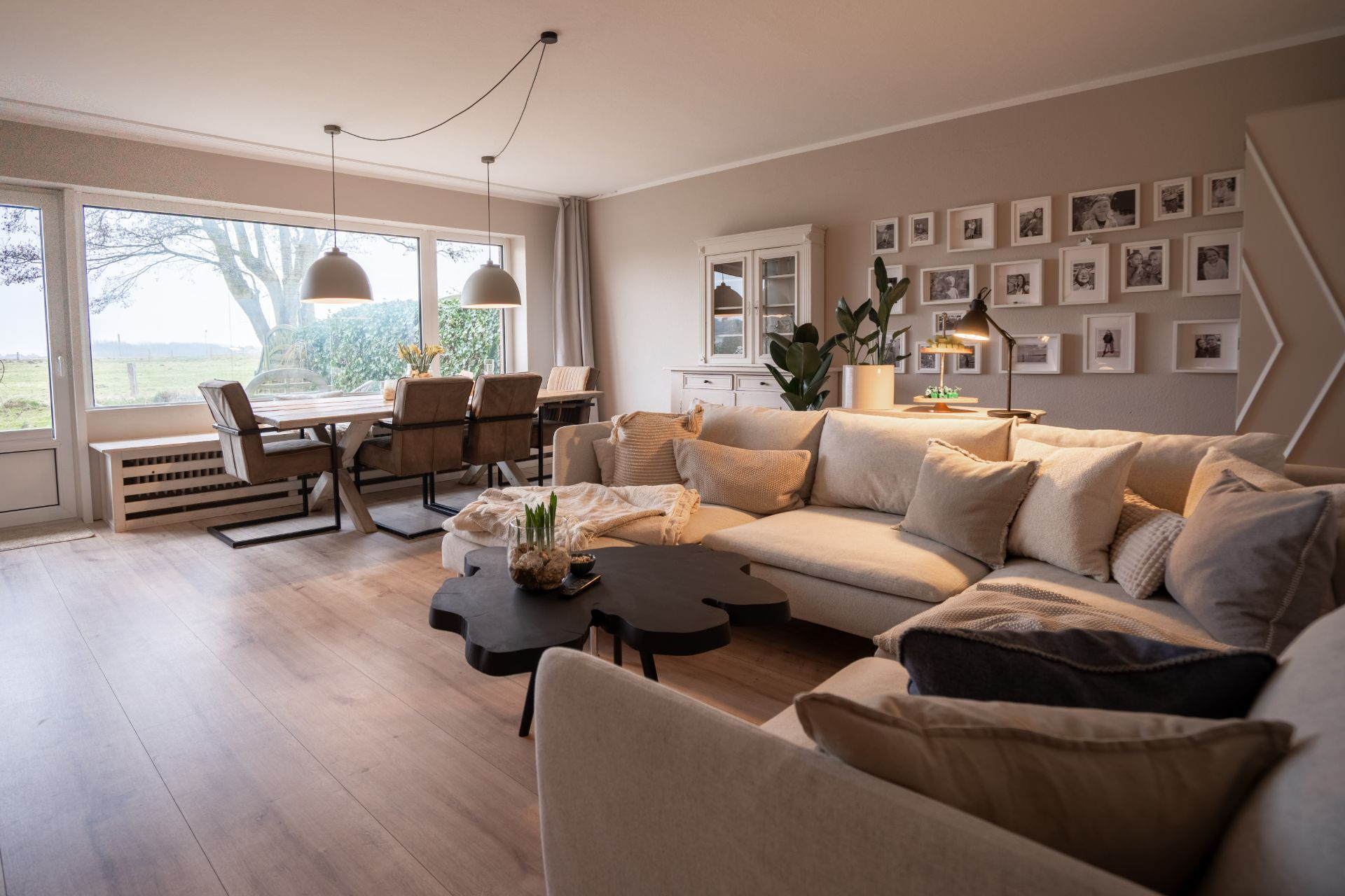 Helles Wohnzimmer mit großer Eckcouch, Esstisch am Fenster und vielen Bildern an der Wand.