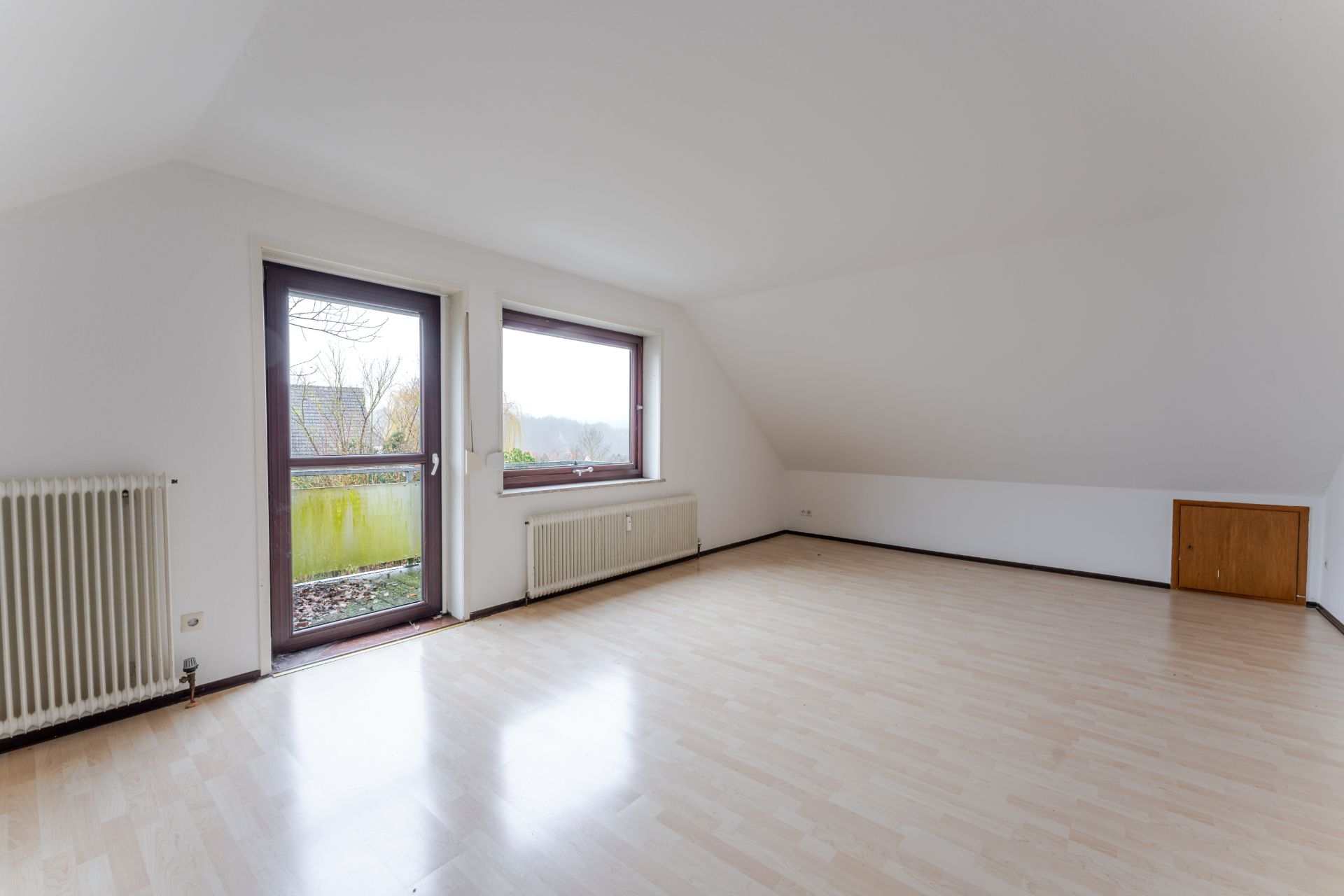 Leeres Zimmer mit hellem Laminatboden, zwei Heizkörpern, Fenster und Tür zum Balkon.