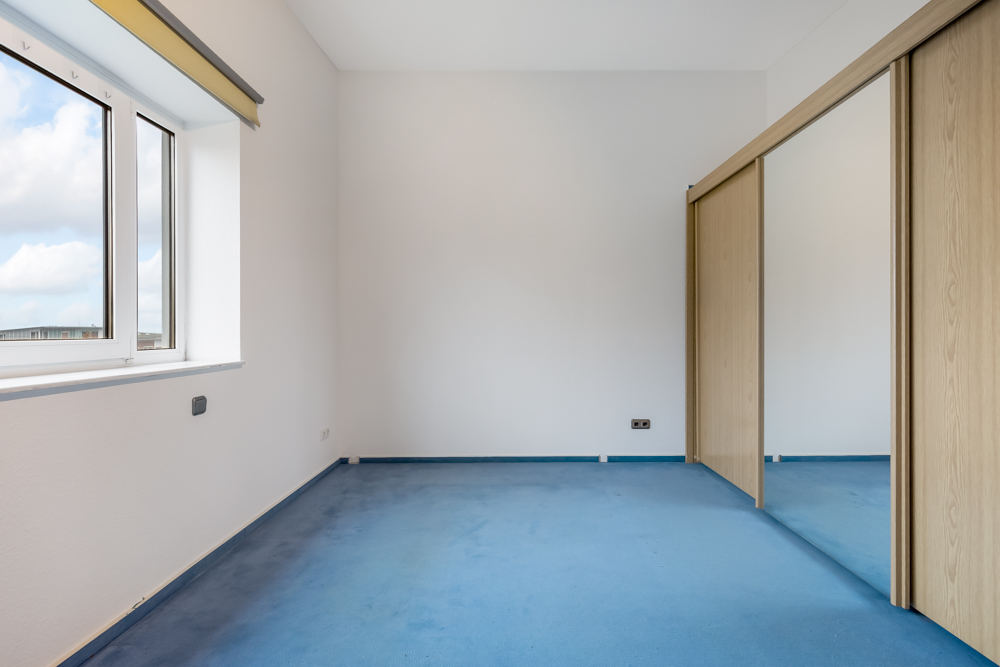 Leeres Zimmer mit blauem Teppich, großem Fenster und Spiegelschrank mit Schiebetüren.