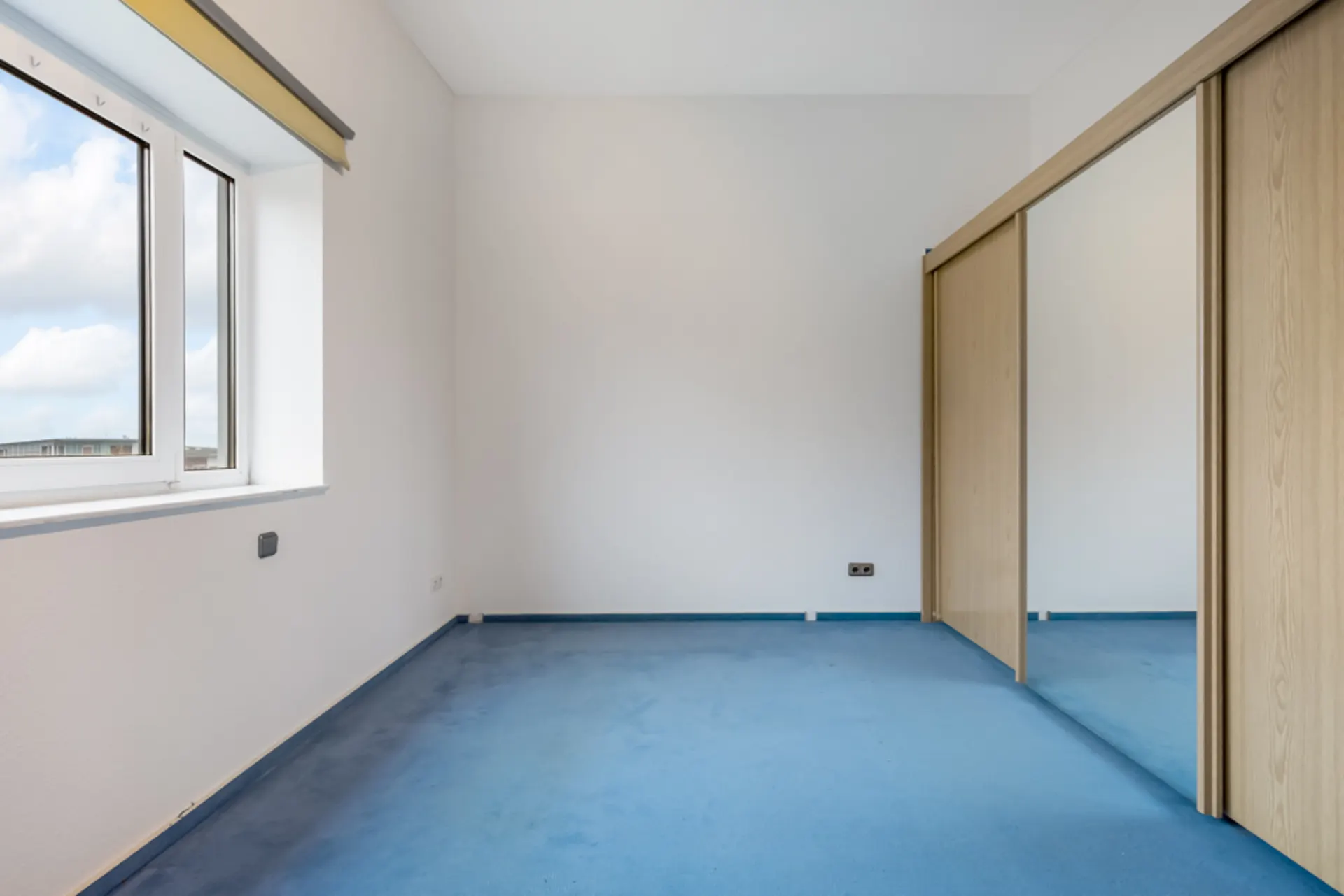 Leeres Zimmer mit blauem Teppich, großem Fenster und Spiegelschrank mit Schiebetüren.