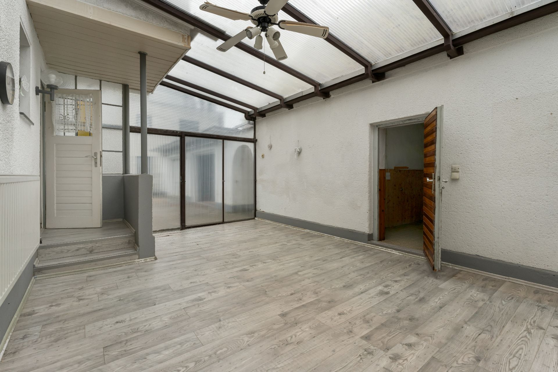 Heller Wintergarten mit Holzboden, Deckenventilator und Türen zu weiteren Räumen.