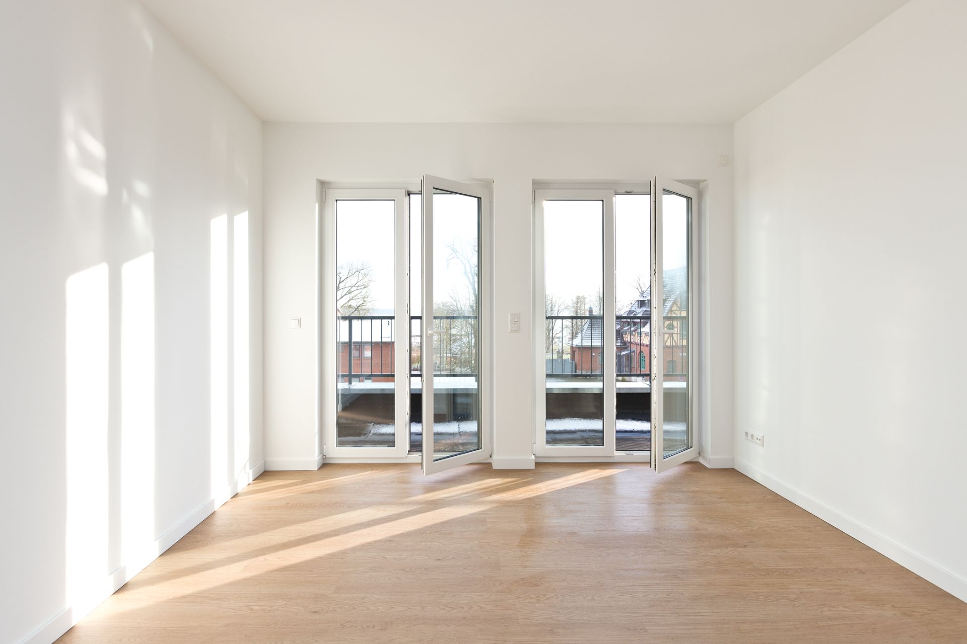 Heller leerer Raum mit Parkettboden und drei großen Fenstertüren zum Balkon.