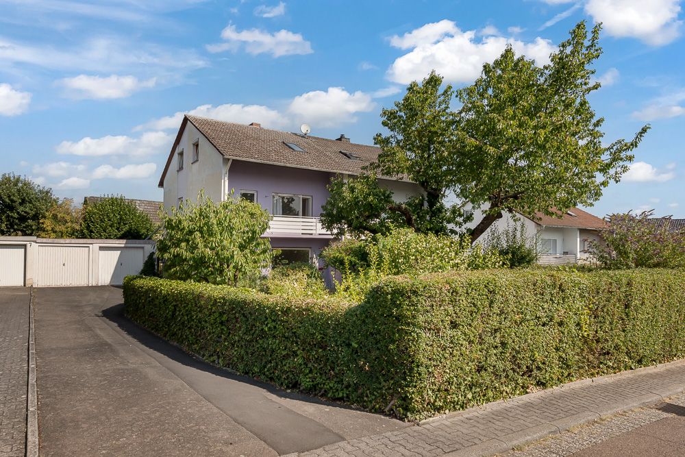 Wohnhaus mit Vorgarten, Hecke und Bäumen an einer Einfahrt, mehrere Garagen im Hintergrund.