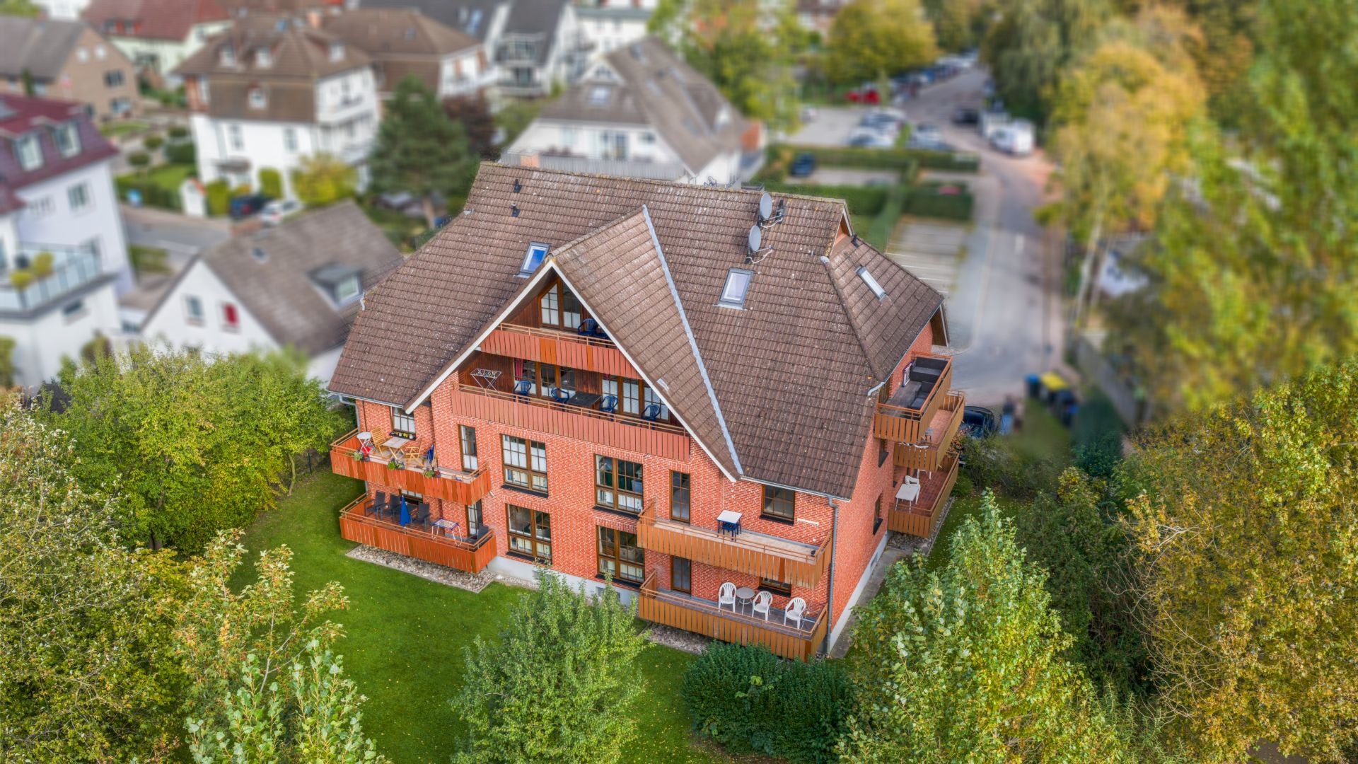 Mehrfamilienhaus mit Balkonen, roter Fassade und Garten, umgeben von Bäumen in Wohngegend.