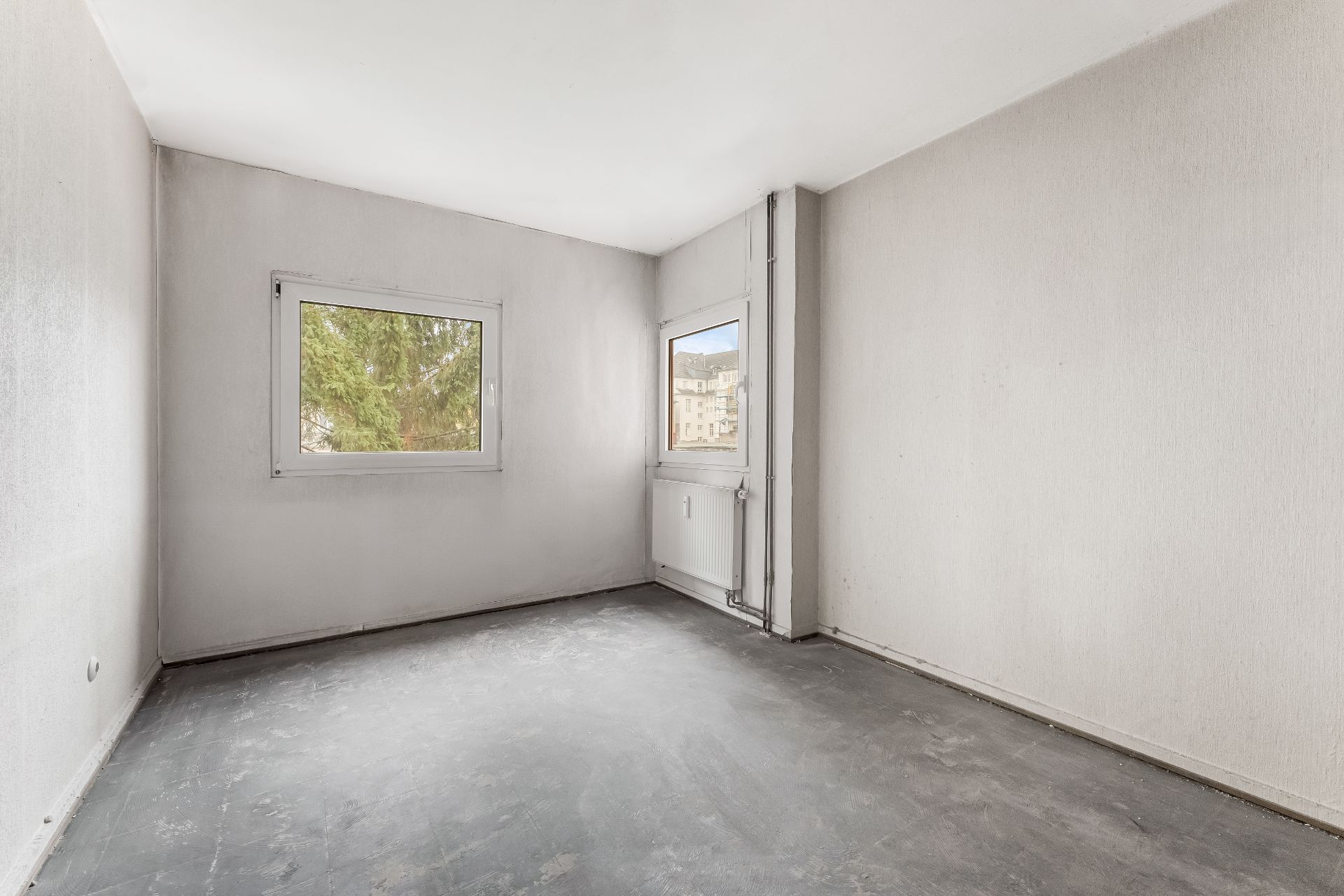 Leeres Zimmer mit zwei Fenstern, grauem Boden und weiß gestrichenen Wänden.