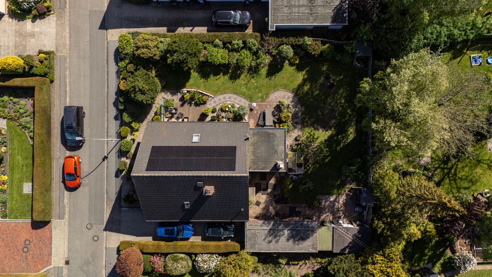 Luftaufnahme eines Hauses mit Garten, Terrasse und parkenden Autos an der Straße.