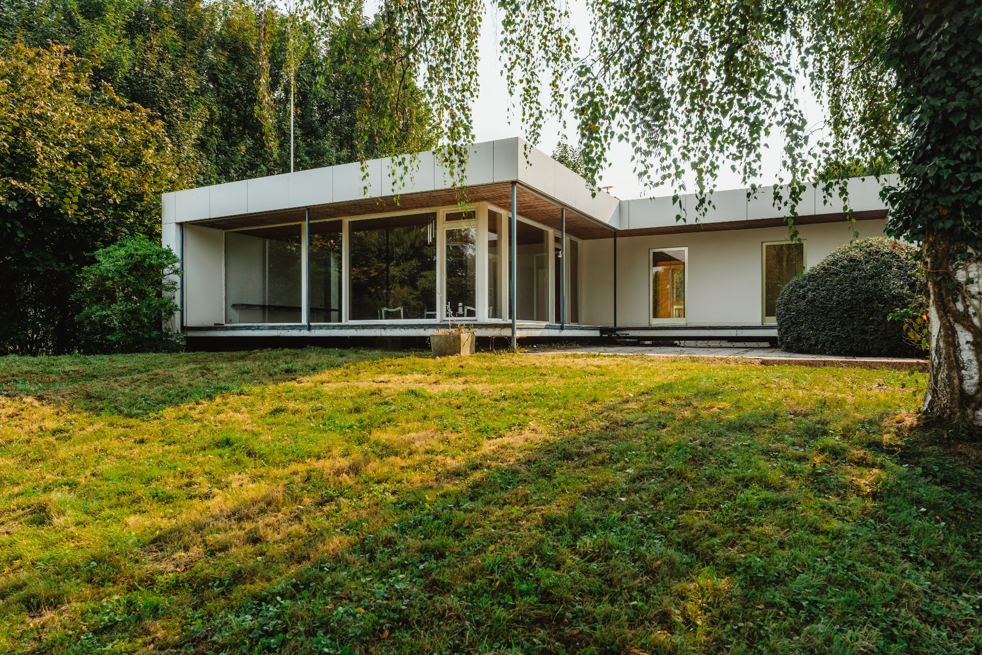Modernes einstöckiges Haus mit großen Fenstern und Terrasse, von Bäumen und Rasen umgeben.
