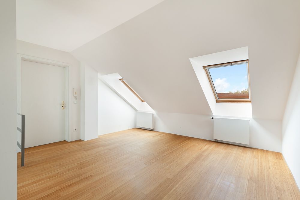 Helles Dachzimmer mit zwei Dachfenstern, Holzboden und weiß gestrichenen Wänden.