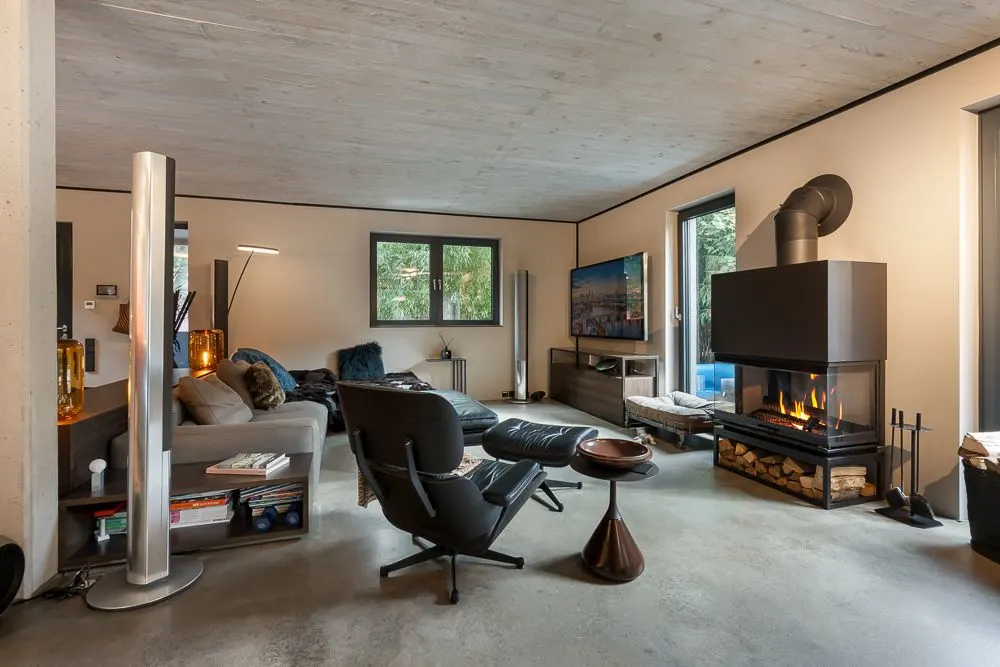 Modernes Wohnzimmer mit Sofa, Sessel, Kamin und großem Fernseher vor Fenstern.