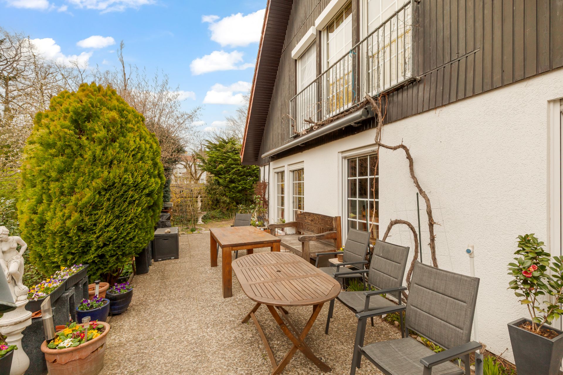 Terrasse mit Holztisch, Stühlen, Pflanzen in Töpfen und angrenzendem Garten an einem Wohnhaus.
