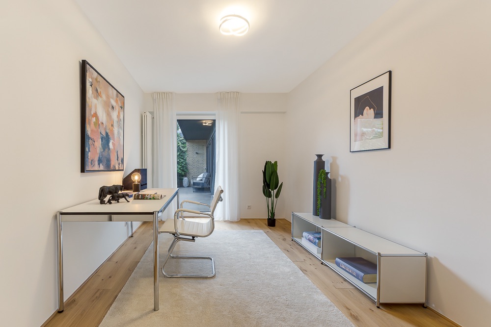 Helles Arbeitszimmer mit Schreibtisch, Stuhl, Regal, Pflanze und Wandbildern.