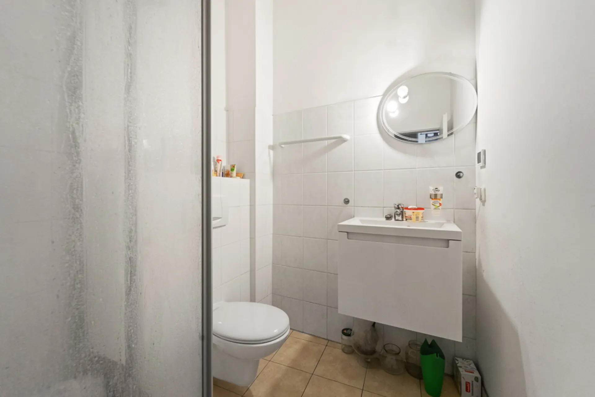 Kleines Bad mit Dusche, Toilette, Waschtisch und ovalem Spiegel an der Wand.