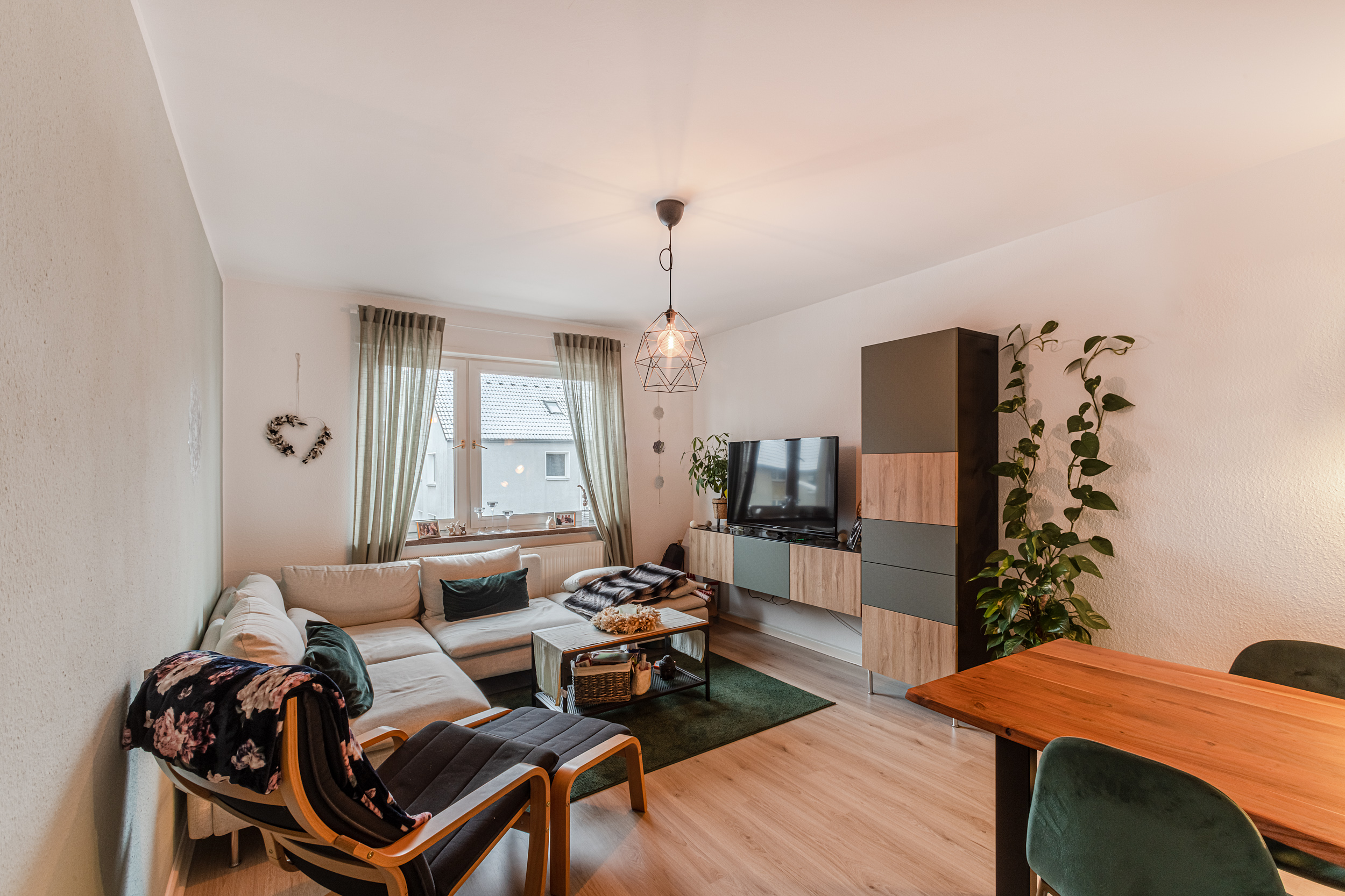 Helles Wohnzimmer mit Ecksofa, Sessel, TV, Esstisch und großen Fenstern mit Vorhängen.
