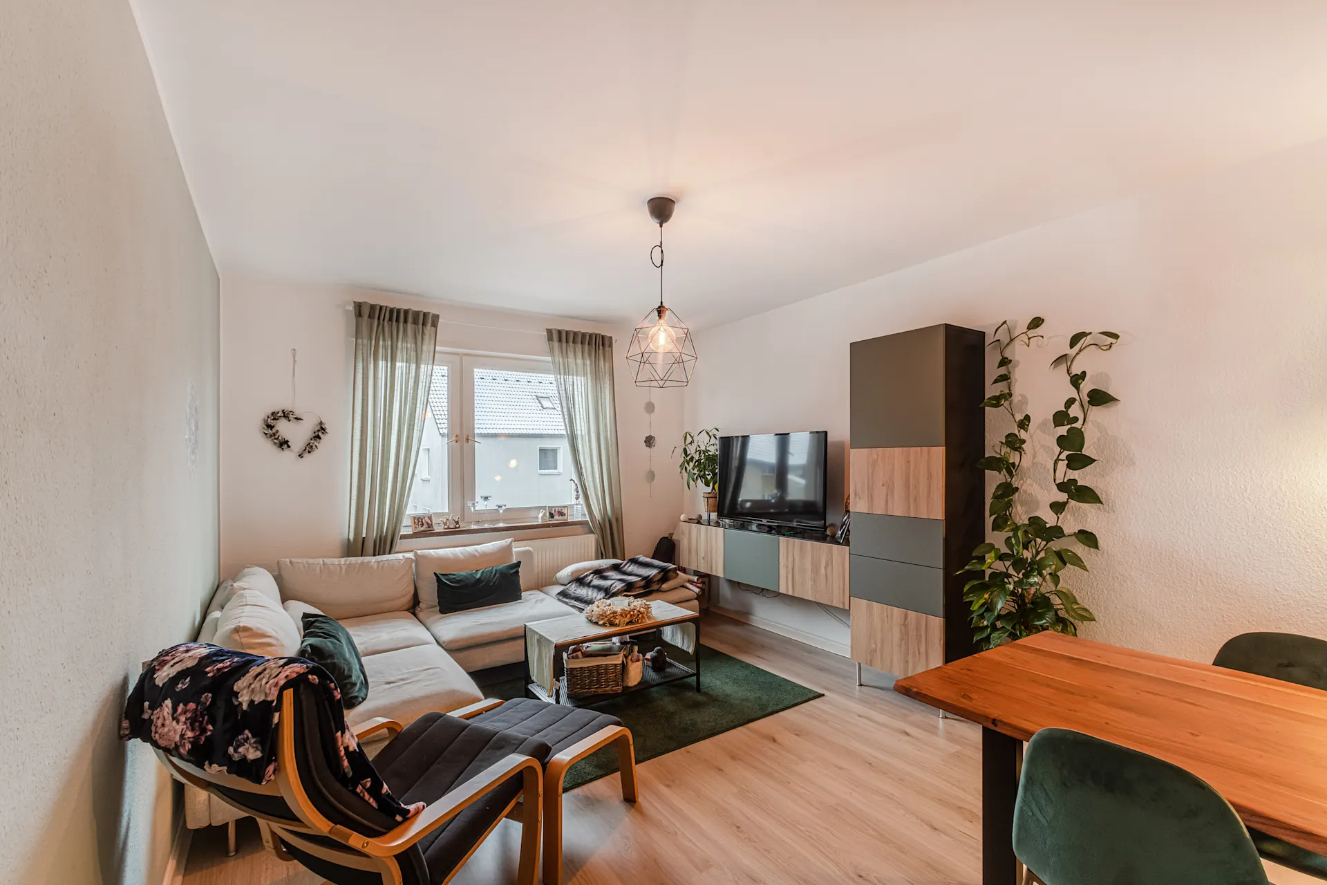Helles Wohnzimmer mit Ecksofa, Sessel, TV, Esstisch und großen Fenstern mit Vorhängen.