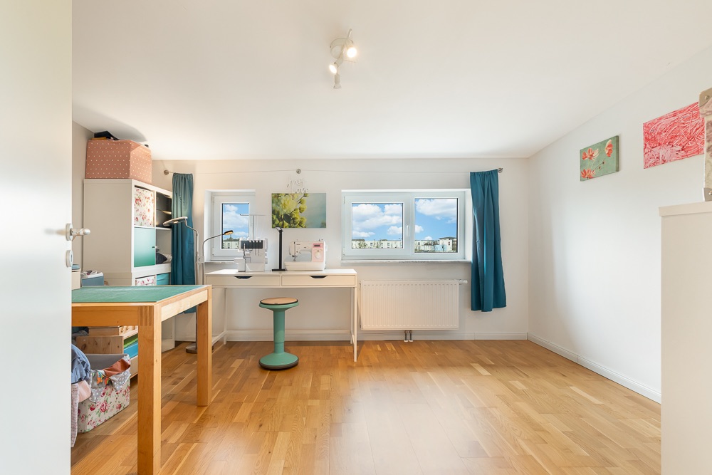 Helles Zimmer mit Holzboden, Schreibtisch, Stuhl, Regalen und mehreren Fenstern.