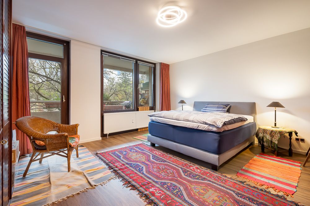 Helles Schlafzimmer mit großem Bett, Teppichen, Korbstuhl und großen Fenstern mit Blick ins Grüne.
