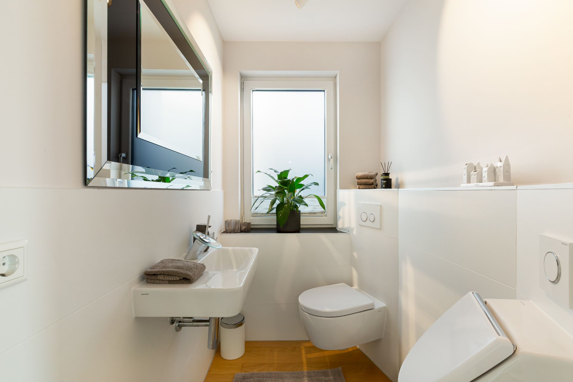 Modernes Badezimmer mit Wandtoilette, Waschbecken, Spiegel und Fenster mit Pflanze.