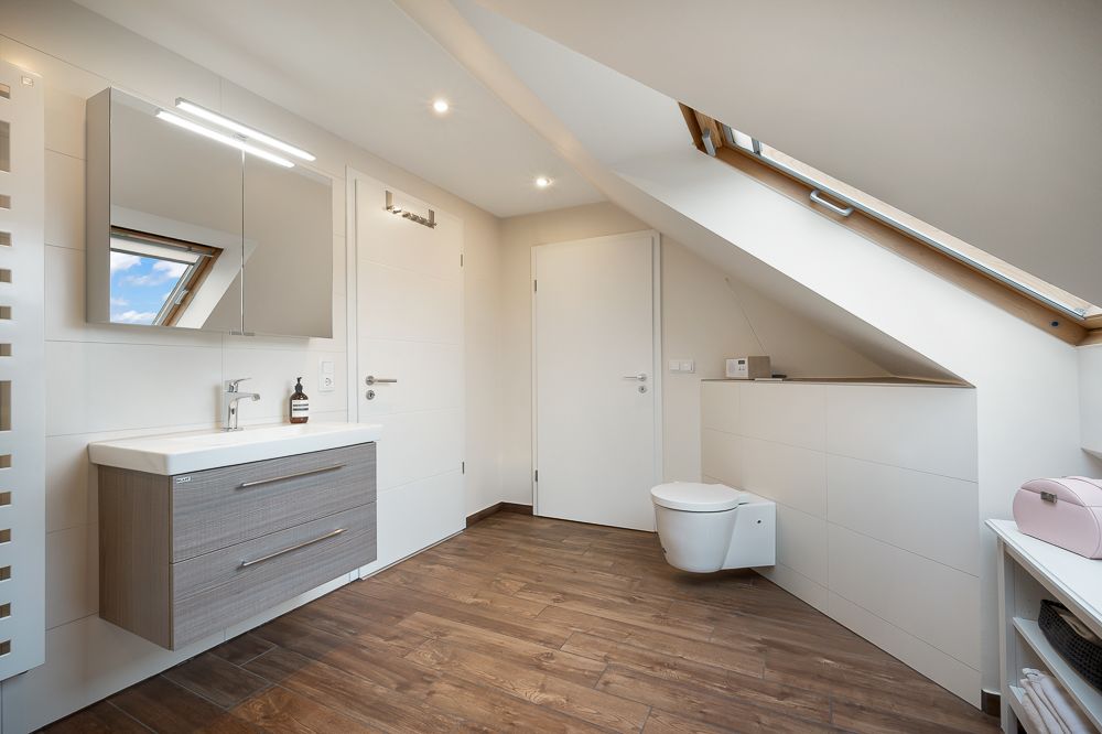 Modernes Badezimmer mit Holzfußboden, Dachfenster, Waschbecken, Spiegel und wandhängender Toilette.