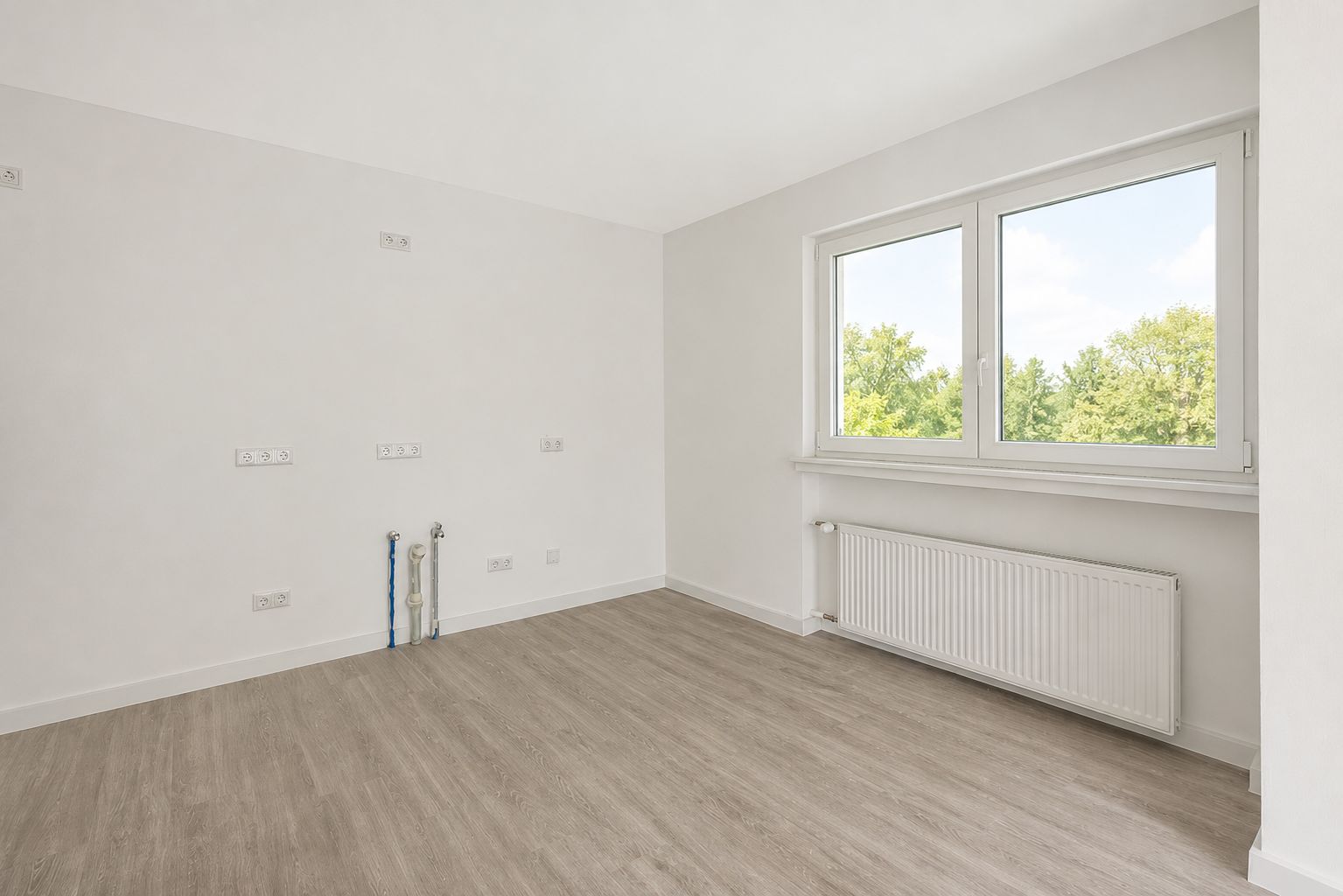 Leerer, heller Raum mit großem Fenster, Heizung und Anschlüssen an der Wand, heller Holzboden.