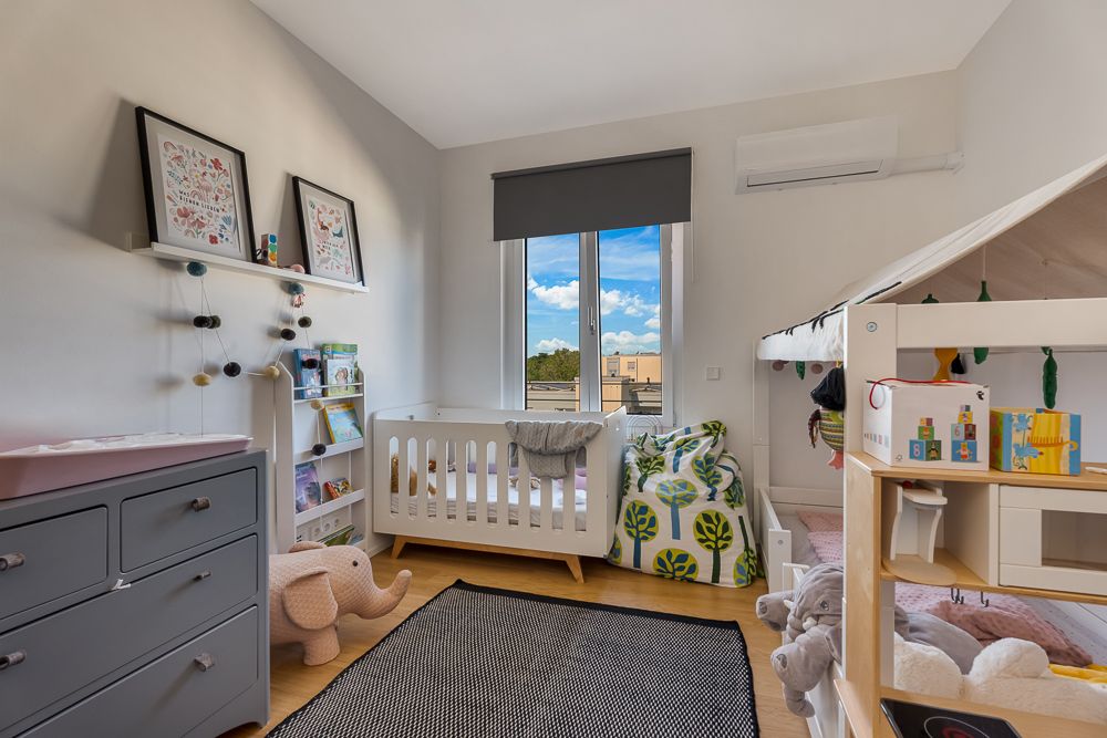 Helles Kinderzimmer mit Babybett, Spielzeug, Regal, Teppich und großem Fenster.