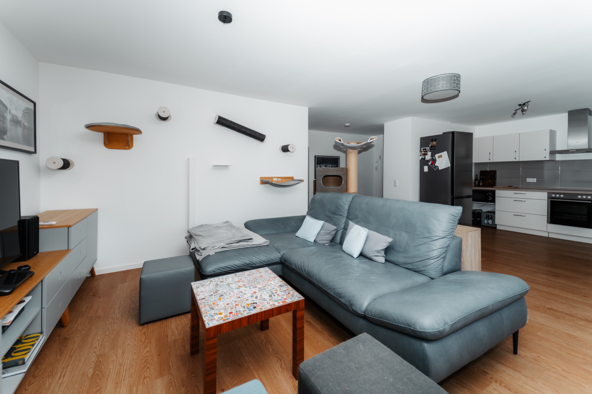 Modernes Wohnzimmer mit grauem Sofa, Fernseher, Holzboden und offener Küche im Hintergrund.