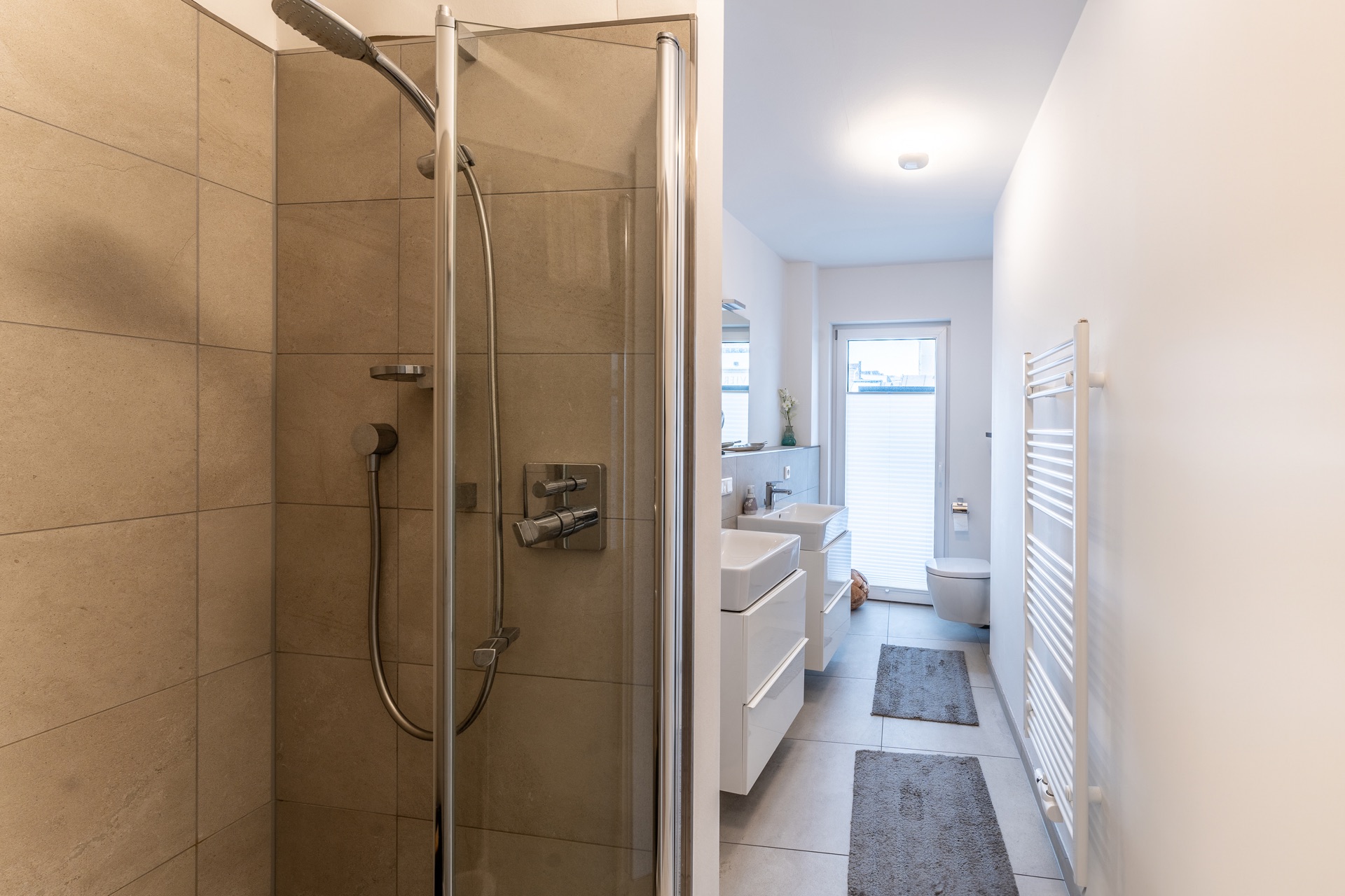 Modernes Badezimmer mit Dusche, Doppelwaschbecken, WC, Heizkörper und zwei Teppichen.