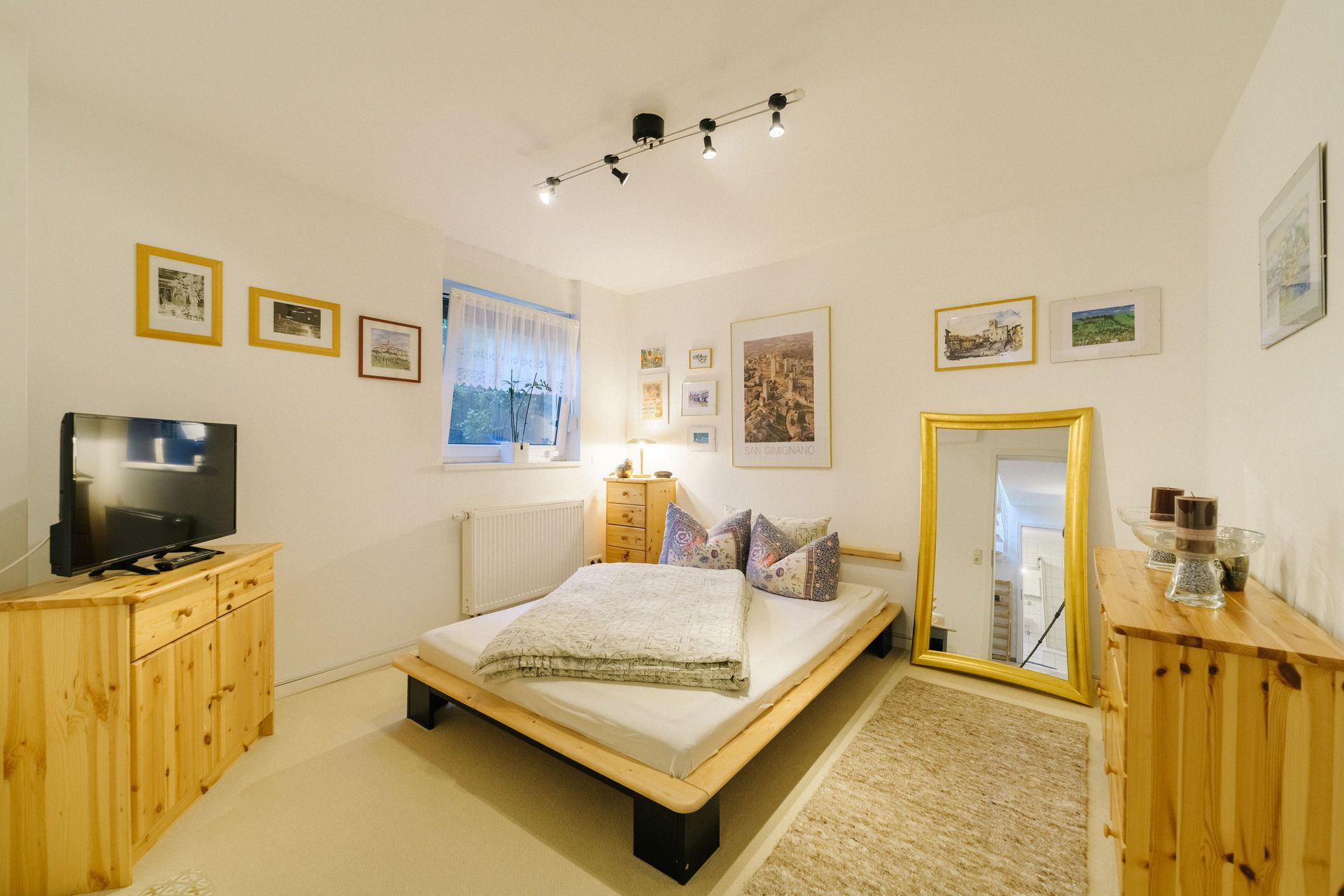 Helles Schlafzimmer mit Doppelbett, Holzkommoden, Spiegel, Fernseher und Bildern an den Wänden.