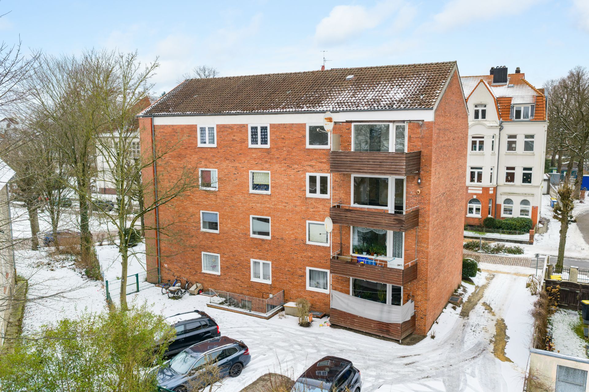 Mehrfamilienhaus aus rotem Backstein mit Balkonen und geparkten Autos im verschneiten Hof.