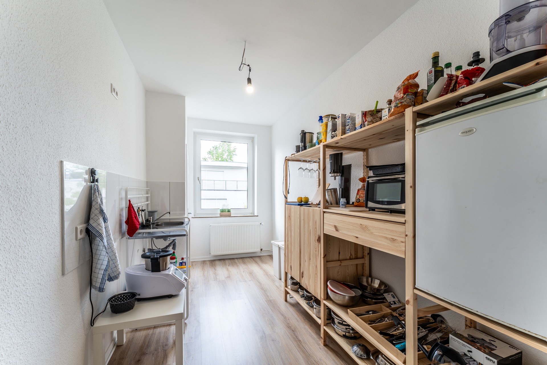 Schmale Küche mit Holzregalen, weißem Kühlschrank und Fenster am Ende des Raums.