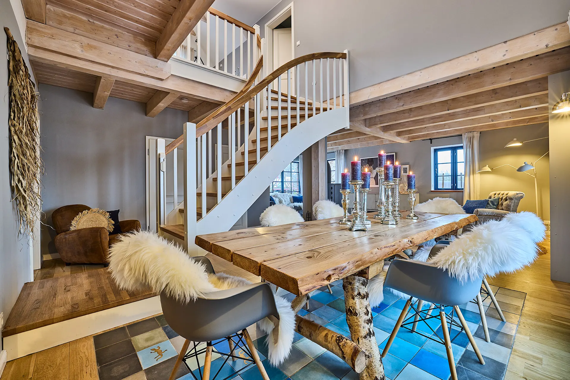 Gemütliches Esszimmer mit Holztisch, Kerzenleuchtern, Fellstühlen und offener Holztreppe.