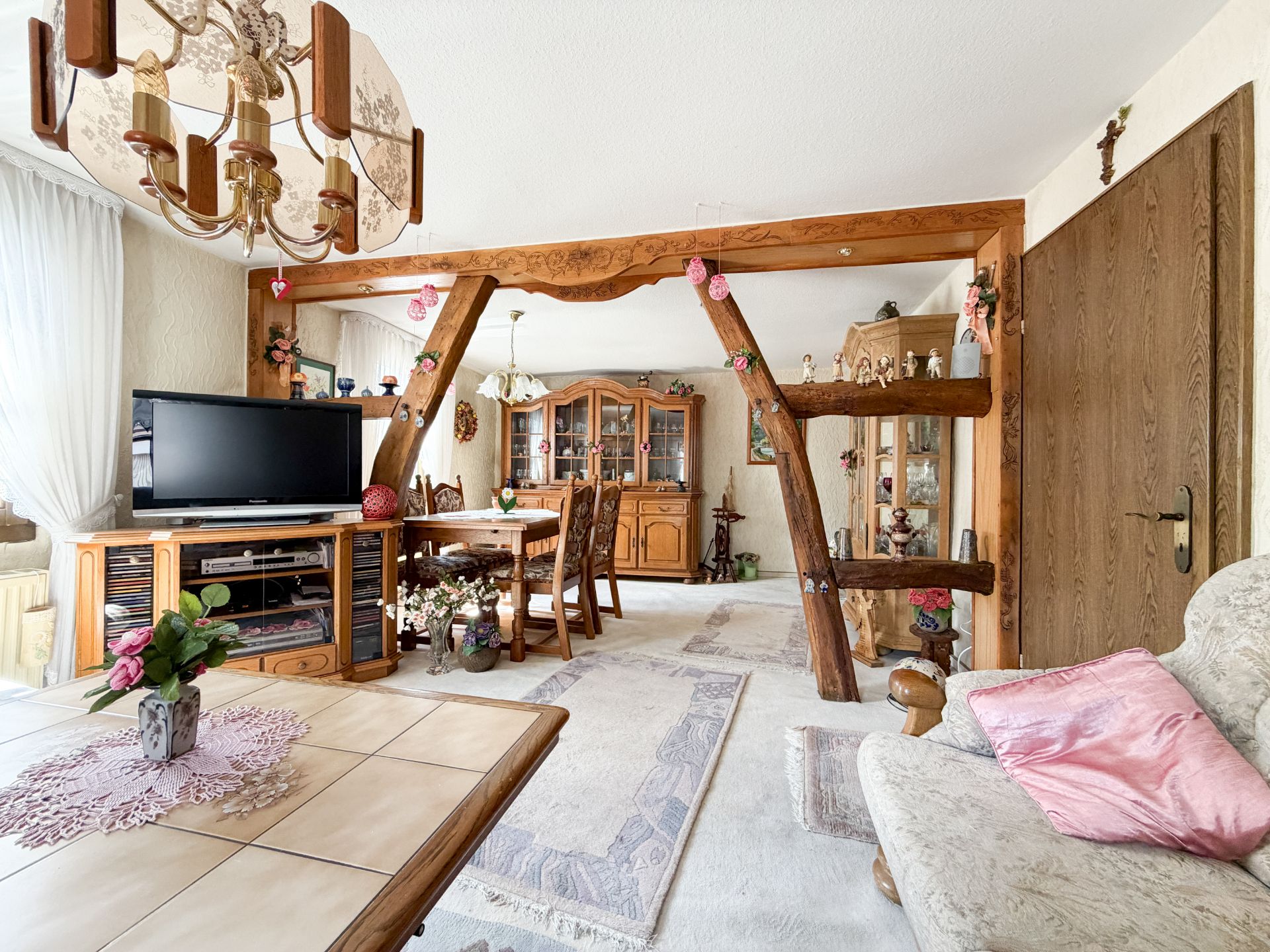 Helles Wohnzimmer mit Holzmöbeln, TV, Esstisch, Sofaecke und großen Fenstern.