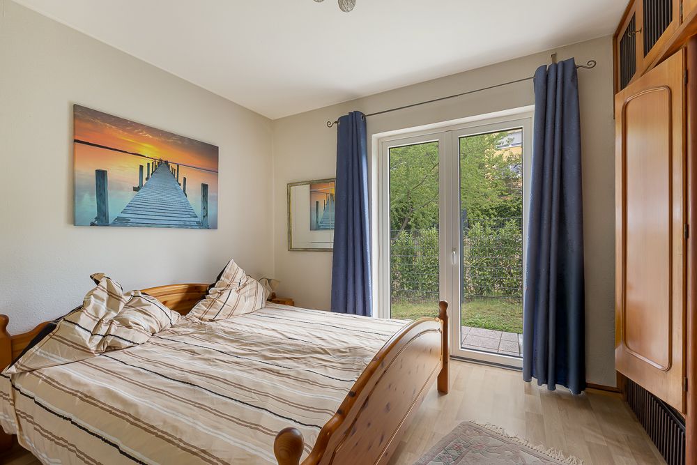 Schlafzimmer mit Holzbett, großem Fenster und blauen Vorhängen, Blick auf den Garten.