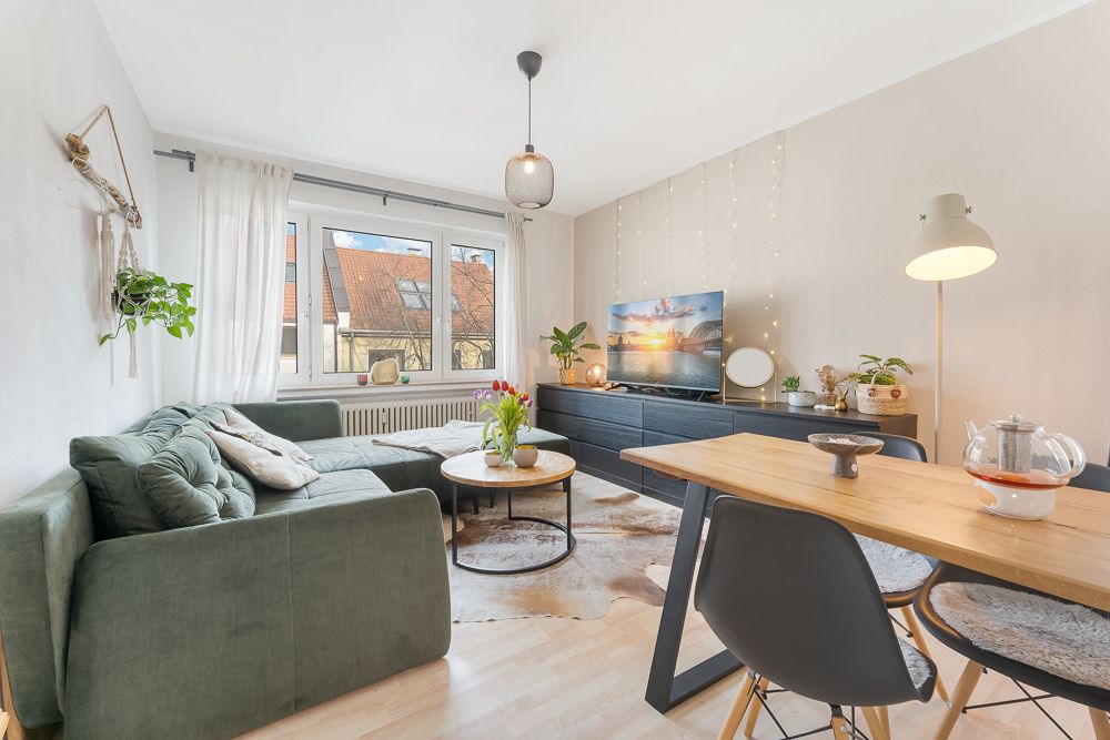 Helles Wohnzimmer mit grünem Sofa, Esstisch, großen Fenstern und Fernseher auf Sideboard.
