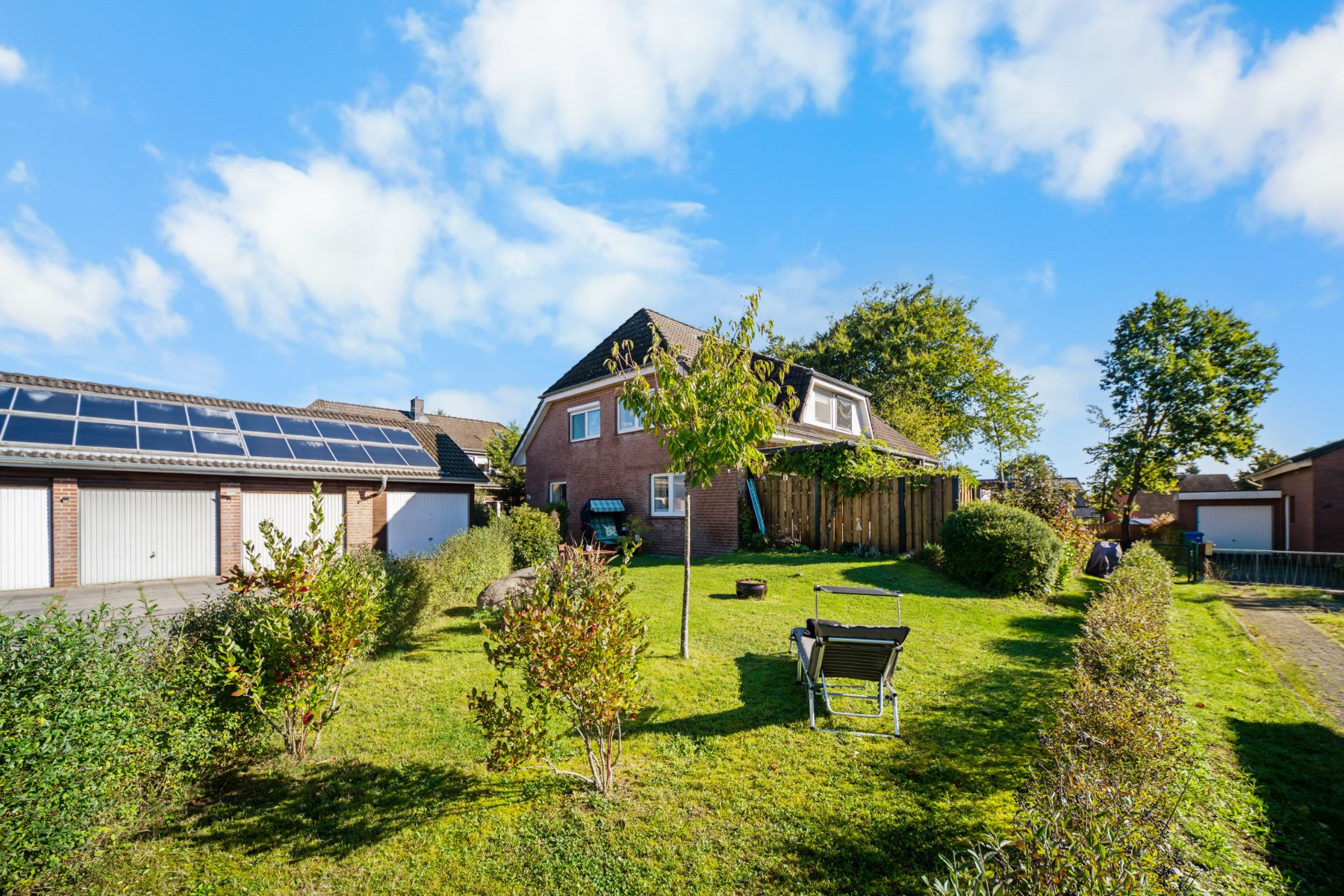 Backsteinwohnhaus mit Garten, Baum, Sträuchern und zwei Gartenstühlen unter blauem Himmel.