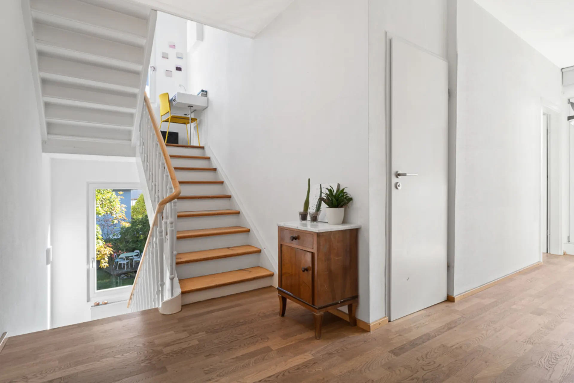 Heller Flur mit Holztreppe, Kommode mit Pflanzen und Fenster mit Blick in den Garten.