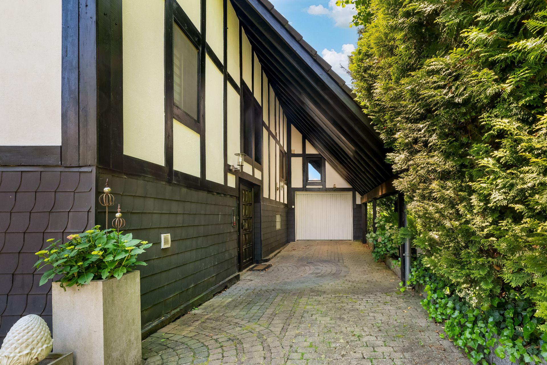 Einfahrt mit Kopfsteinpflaster, Fachwerkhaus, Garage und grünen Büschen am Rand.