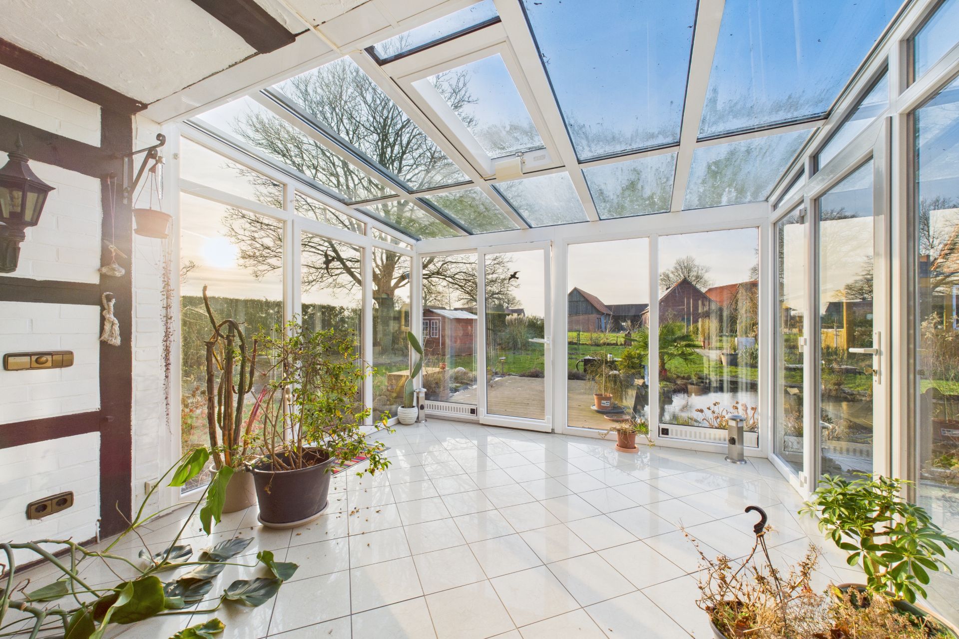 Heller Wintergarten mit vielen Fenstern, Fliesenboden und einigen Topfpflanzen, Blick in den Garten.