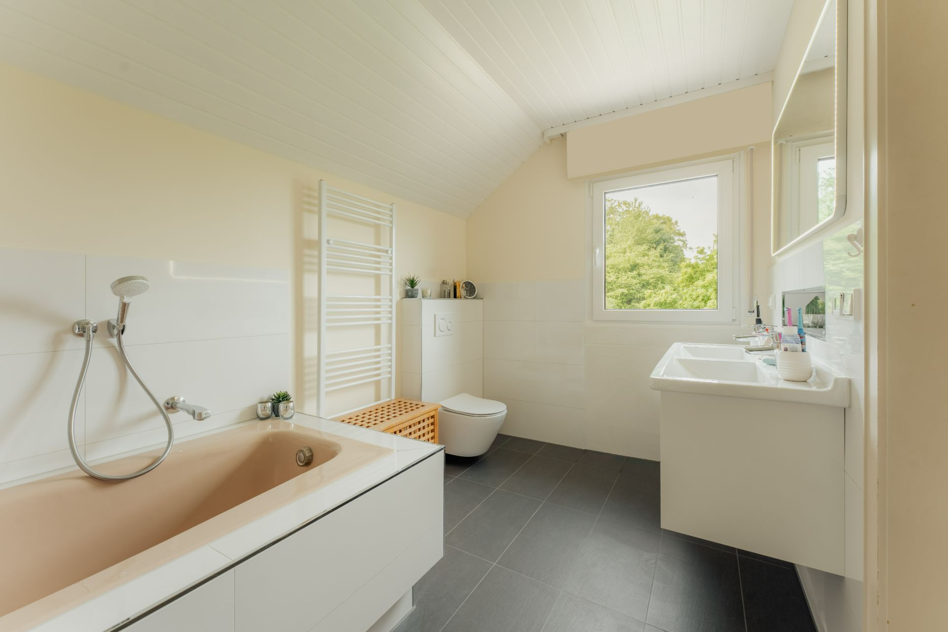 Helles Badezimmer mit Badewanne, Toilette, Waschbecken und Fenster mit Blick ins Grüne.