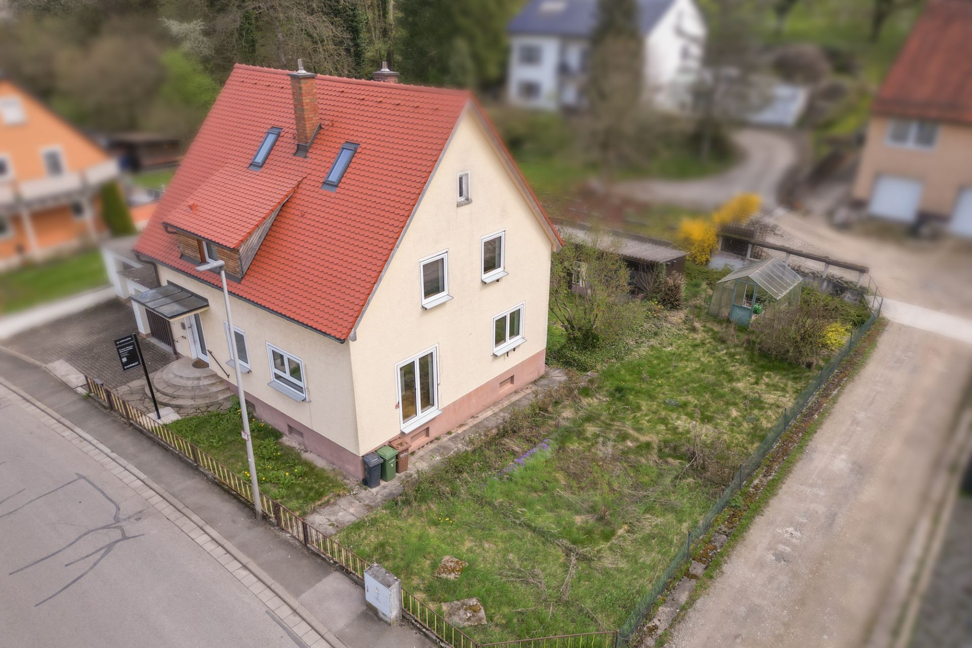 Zweistöckiges Haus mit rotem Dach und Garten, von einem Zaun umgeben, an einer Straßenecke gelegen.