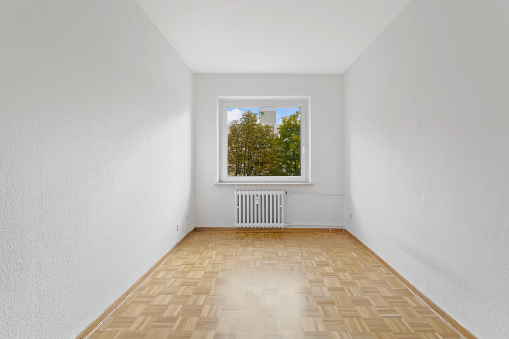 Leerer, heller Raum mit Parkettboden, weißem Heizkörper und Fenster mit Blick auf Bäume.